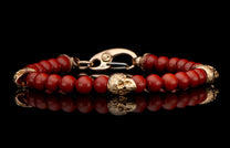 Red jasper beaded bracelet with an 18K gold skull centerpiece, ornate end caps, and carabiner clasp, reflected.