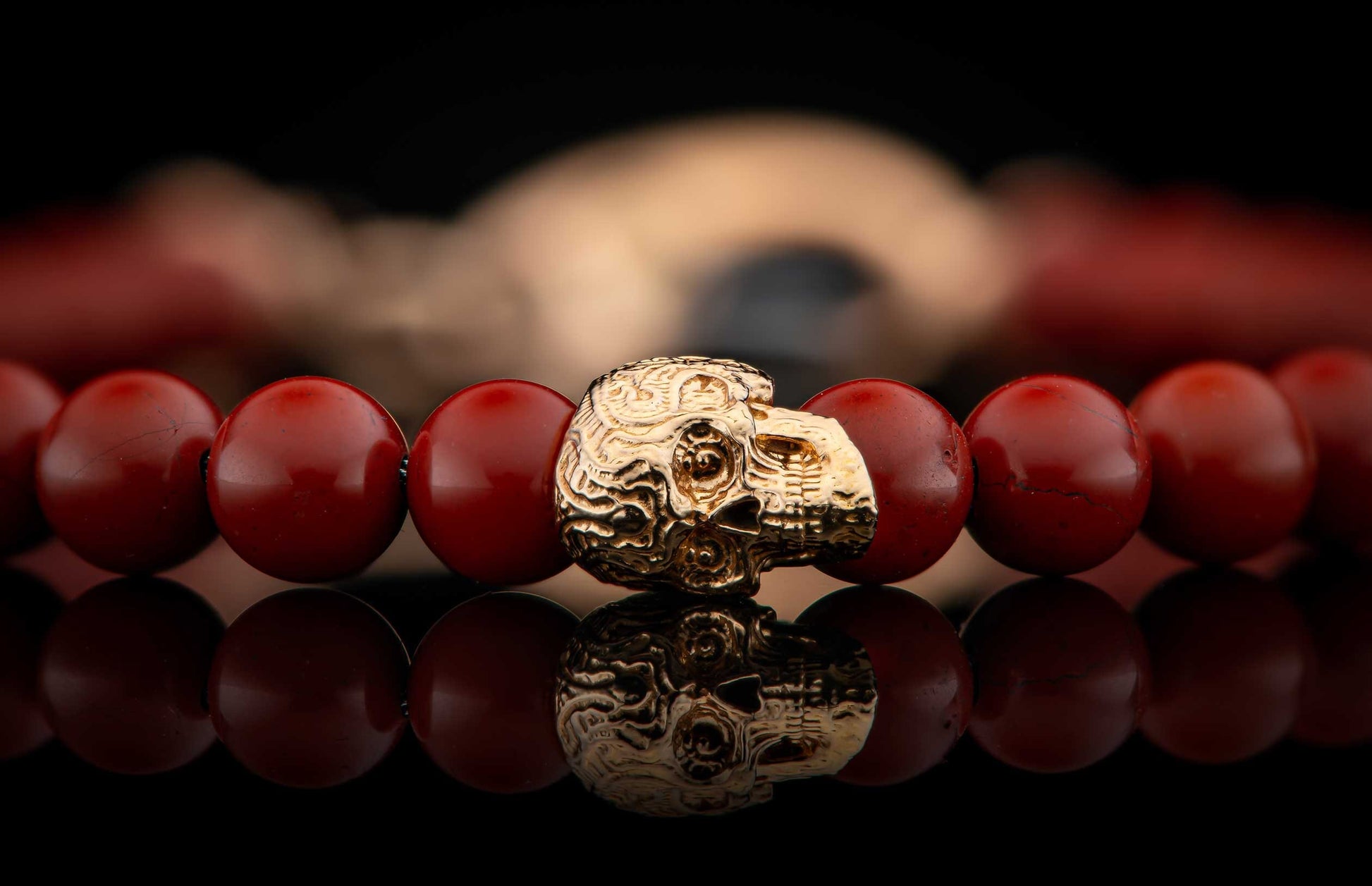 Red beaded bracelet featuring a detailed gold skull charm, reflected on a dark surface.