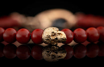 Red beaded bracelet featuring a detailed gold skull charm, reflected on a dark surface.