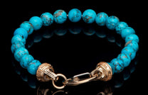 Turquoise beaded bracelet with natural matrix, featuring a gold-toned clasp with engraved details, reflected on a dark surface.