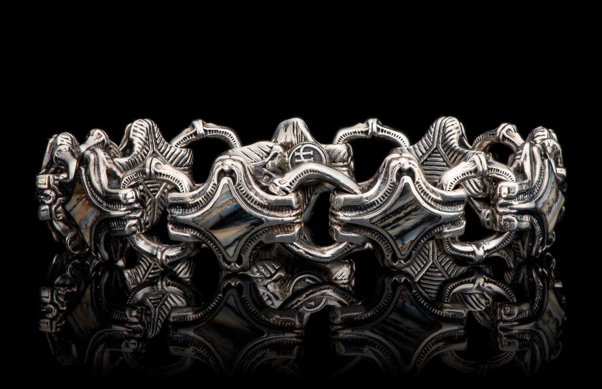 Silver bracelet with intricate, sculptural links and diamond-shaped inlays, reflecting on a black surface.