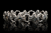Silver bracelet with intricate, sculptural links and diamond-shaped inlays, reflecting on a black surface.