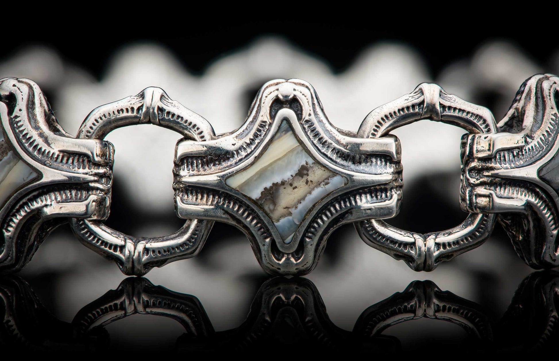 Ornate silver-toned link bracelet with textured details and mother-of-pearl inlay, reflecting on a dark surface.