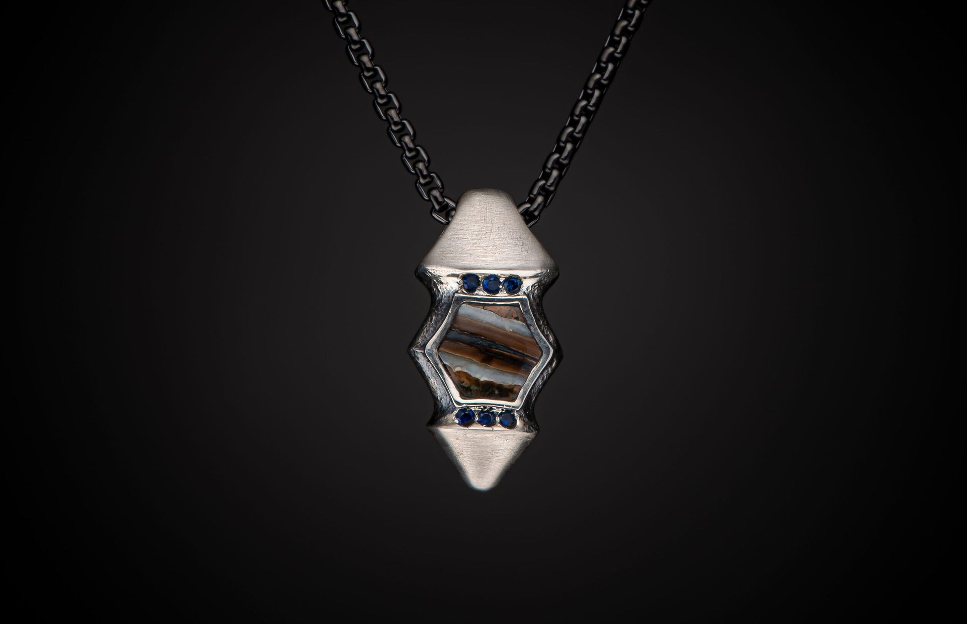 Silver pendant featuring a hexagonal fossilized stone, accented with blue sapphires, suspended on a dark chain.