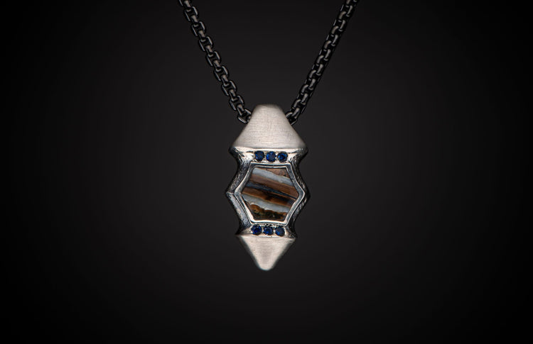 Silver pendant featuring a hexagonal fossilized stone, accented with blue sapphires, suspended on a dark chain.