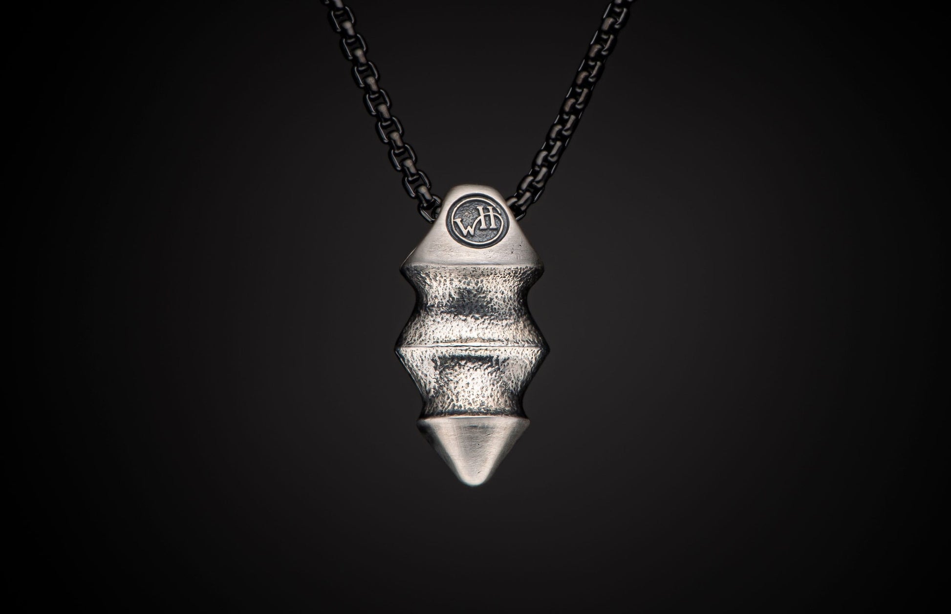 Silver-toned textured pendant displaying a 'WH' logo, geometric ridged body, pointed tip, and a dark chain.