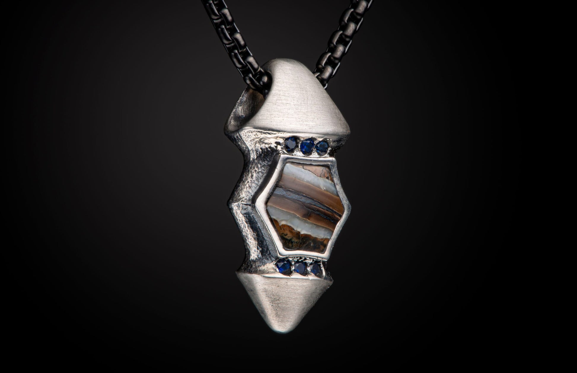 Textured silver pendant with a hexagonal brown and white striated stone, accented by six small blue gemstones, on a dark chain.