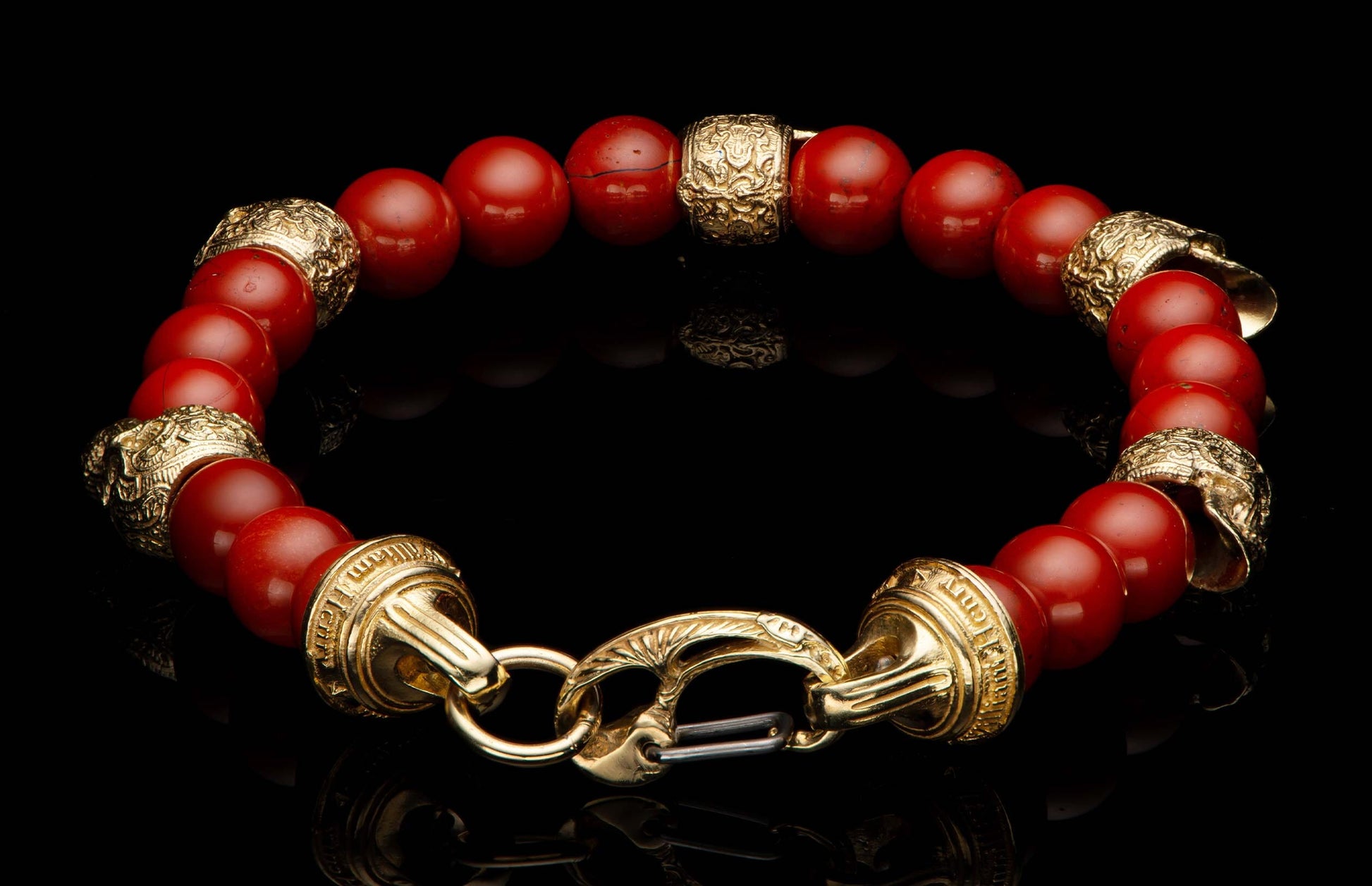 Red bead bracelet with ornate gold-toned metal spacers and a decorative clasp on a reflective black surface.
