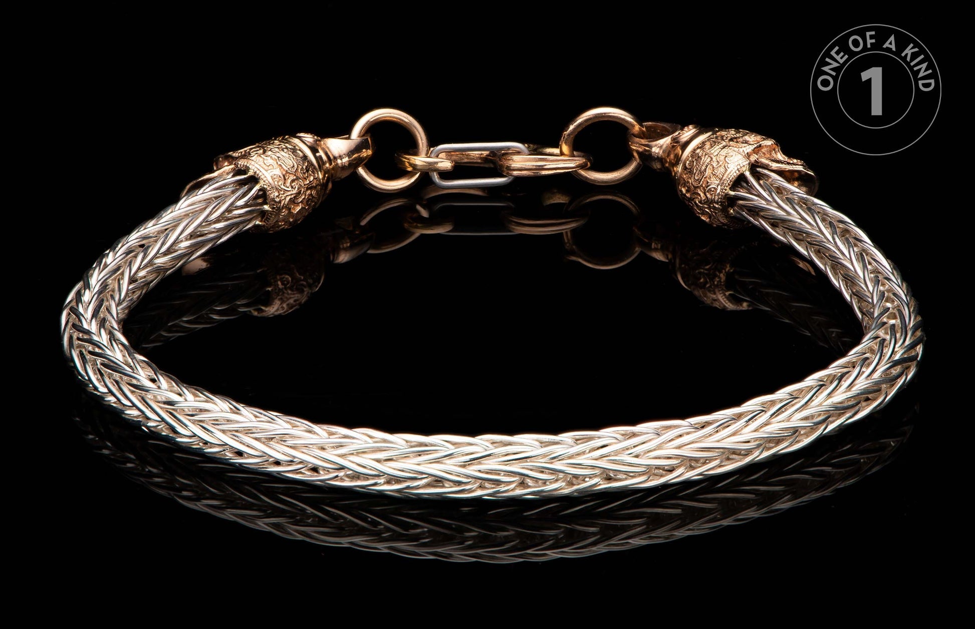 Braided silver-tone metal bracelet with ornate gold-tone caps and clasp, reflecting on a black surface, with a 'One of a Kind' label.
