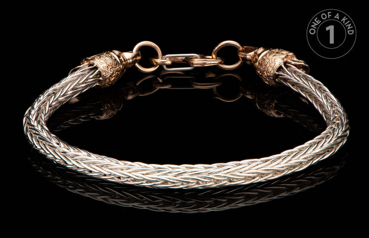 Braided silver-tone metal bracelet with ornate gold-tone caps and clasp, reflecting on a black surface, with a 'One of a Kind' label.