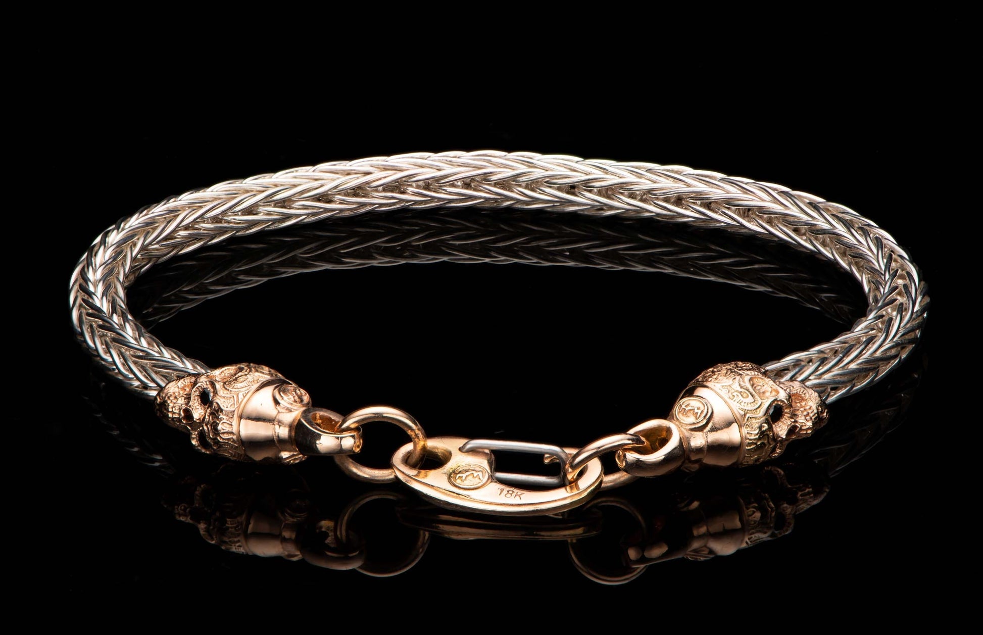 Braided silver metal bracelet with rose gold skull ends and an 18K marked clasp, reflected on black.
