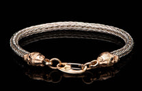 Braided silver metal bracelet with rose gold skull ends and an 18K marked clasp, reflected on black.
