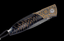 Folding knife with dark handle featuring warm organic patterns, decorative bolster with gold sun and star engravings, and a Damascus steel blade.