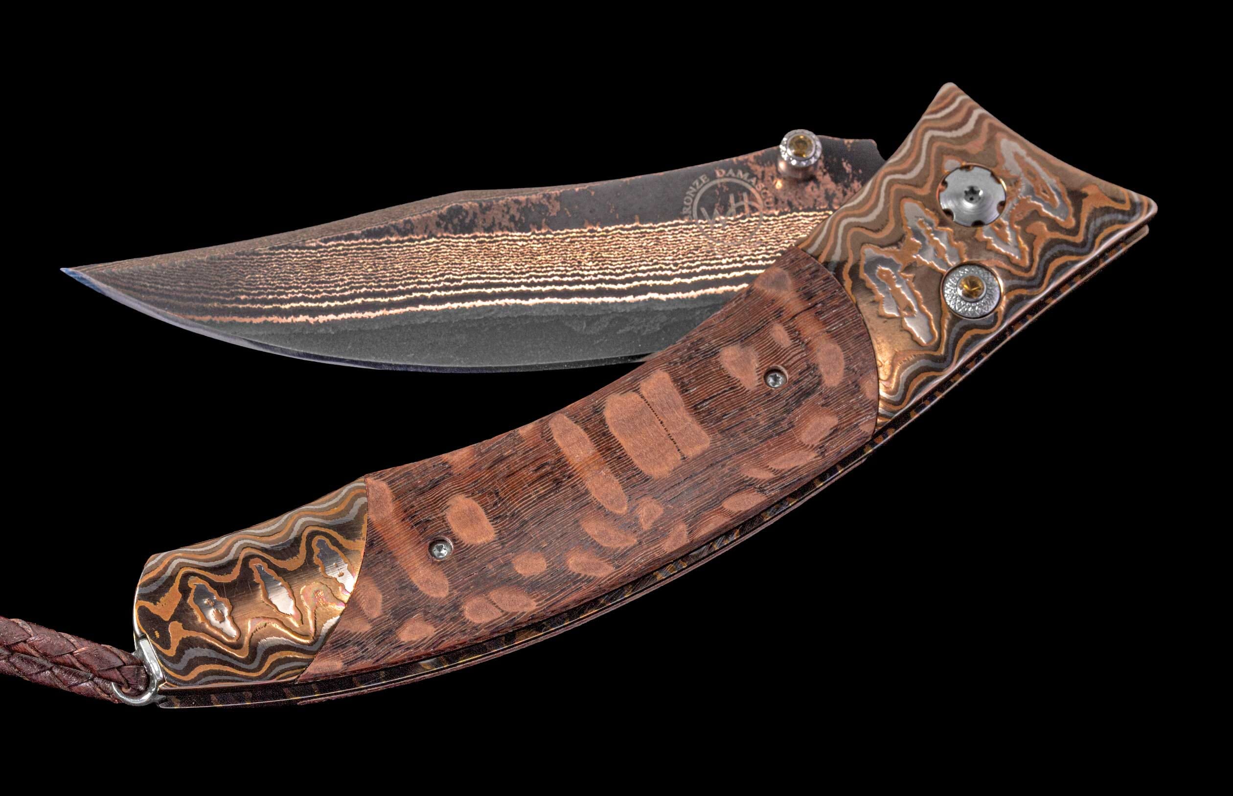 Open folding knife with a wavy bronze Damascus blade showing 'BRONZE DAMASCUS' text, a spotted reddish-brown wood handle, and ornate Damascus bolsters with yellow jewels.