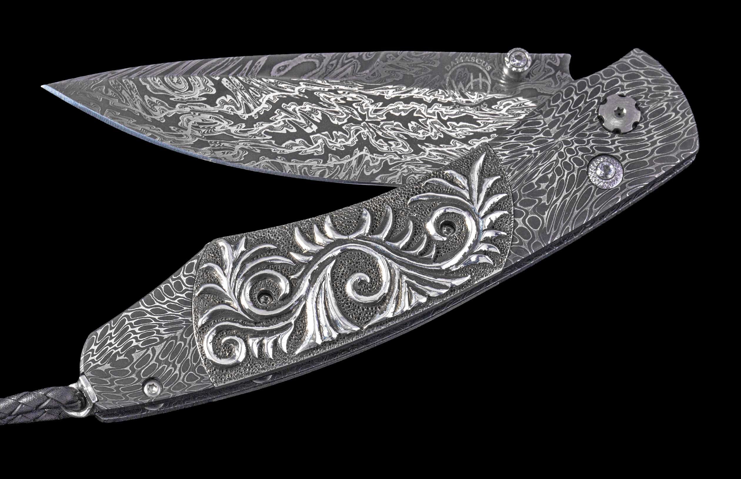 Open Damascus steel folding knife with intricate patterns, an ornate silver scrollwork handle inlay, gemstones, and a braided leather lanyard.