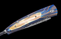 Blue, gold, and silver wave-patterned knife with mother-of-pearl inlay, decorative screws, and a black braided lanyard.