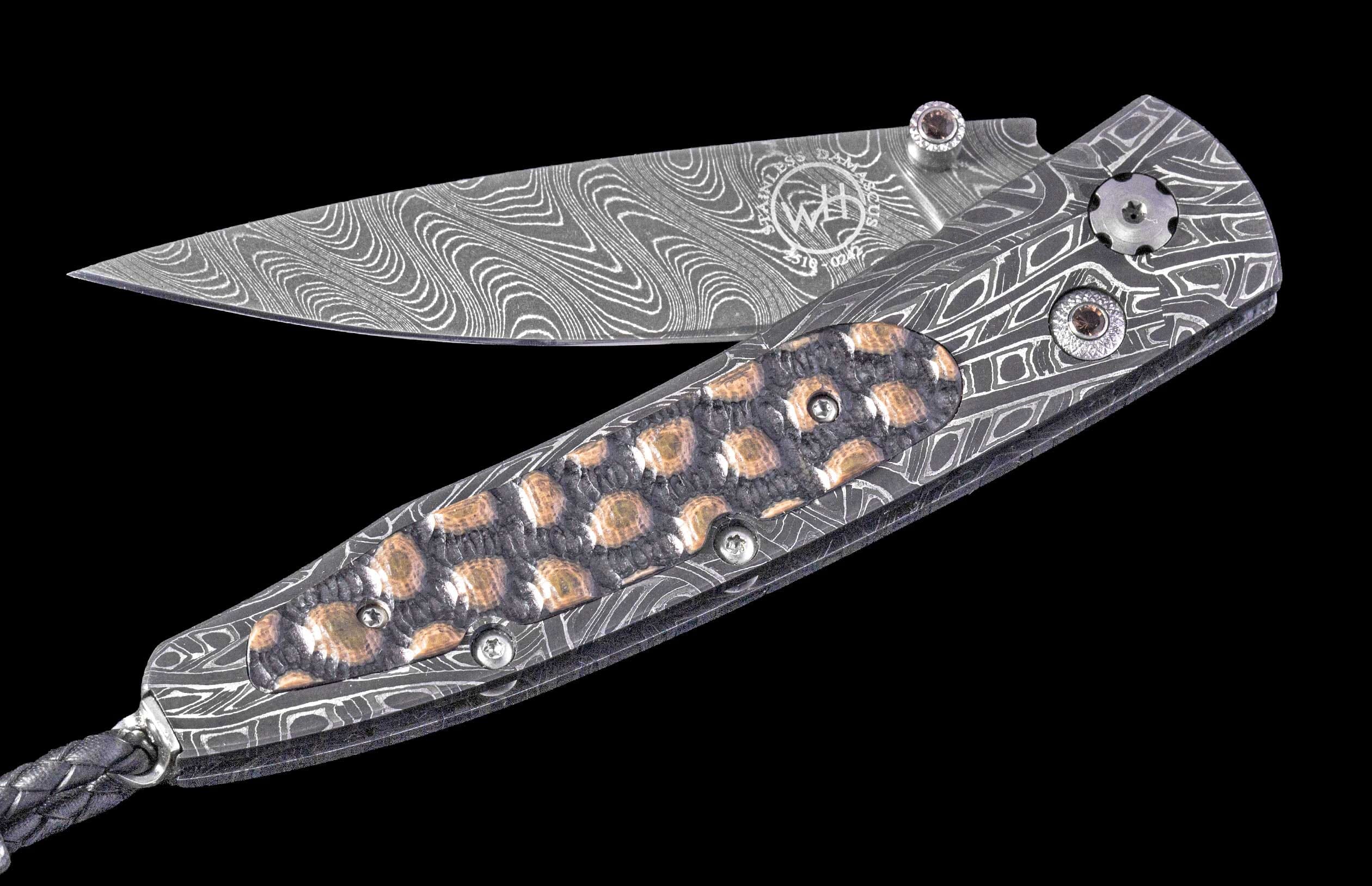 Damascus steel folding knife with wavy patterns, a textured handle with gold and gem accents, and a braided lanyard.