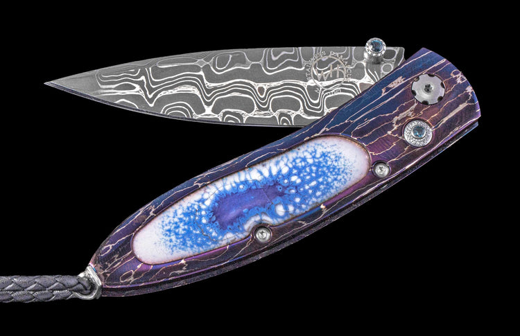 Folding knife with a wavy patterned Damascus steel blade, an iridescent purple handle featuring a cracked white and blue inlay, and blue gem accents.