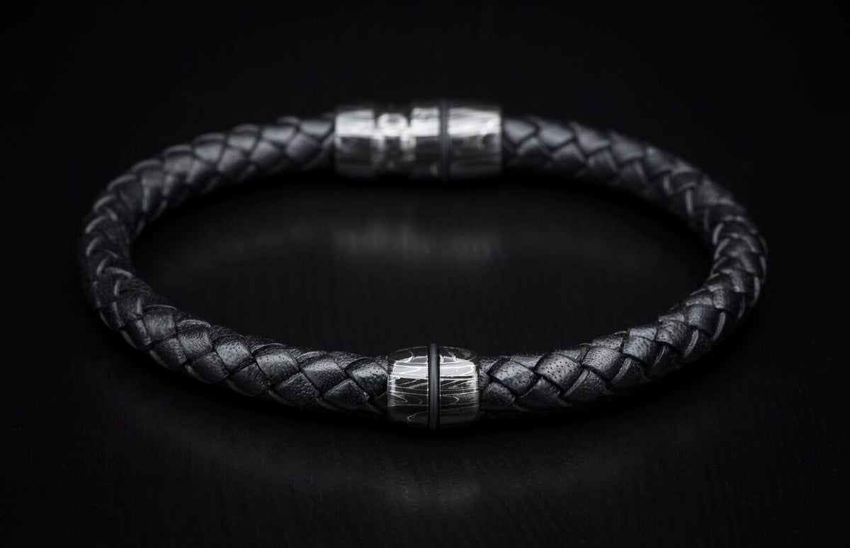 Black braided leather bracelet with a silver metal bead featuring a wavy pattern and a blurred magnetic clasp.