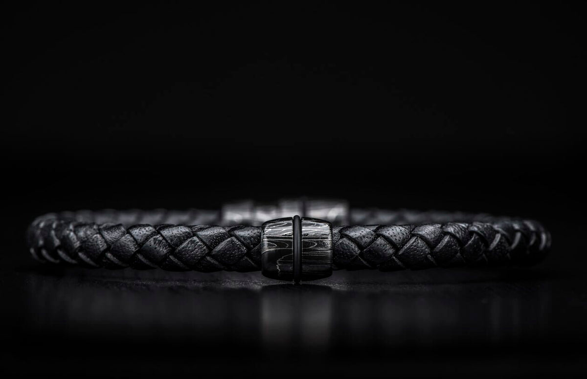 Black braided leather bracelet with a dark metal clasp featuring a distinct wavy pattern, centered.