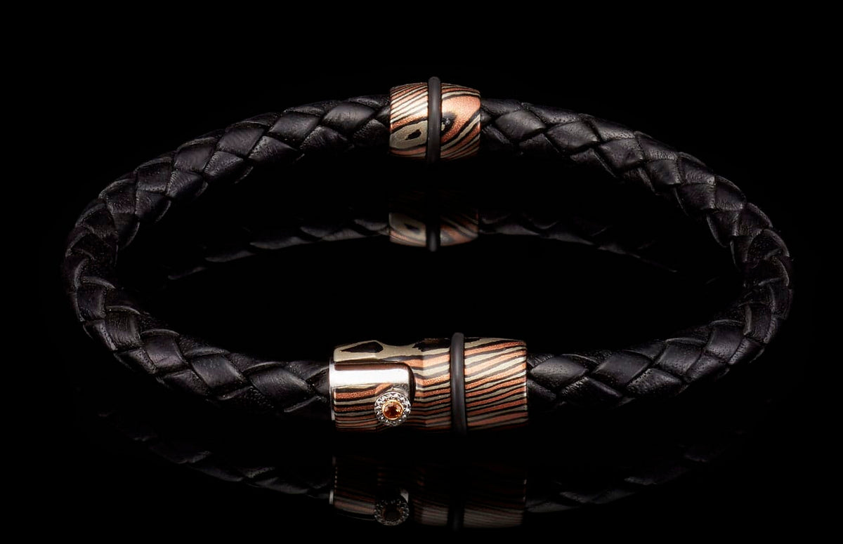 Black braided leather bracelet with two copper and silver patterned metal beads, one accented with an orange gemstone.
