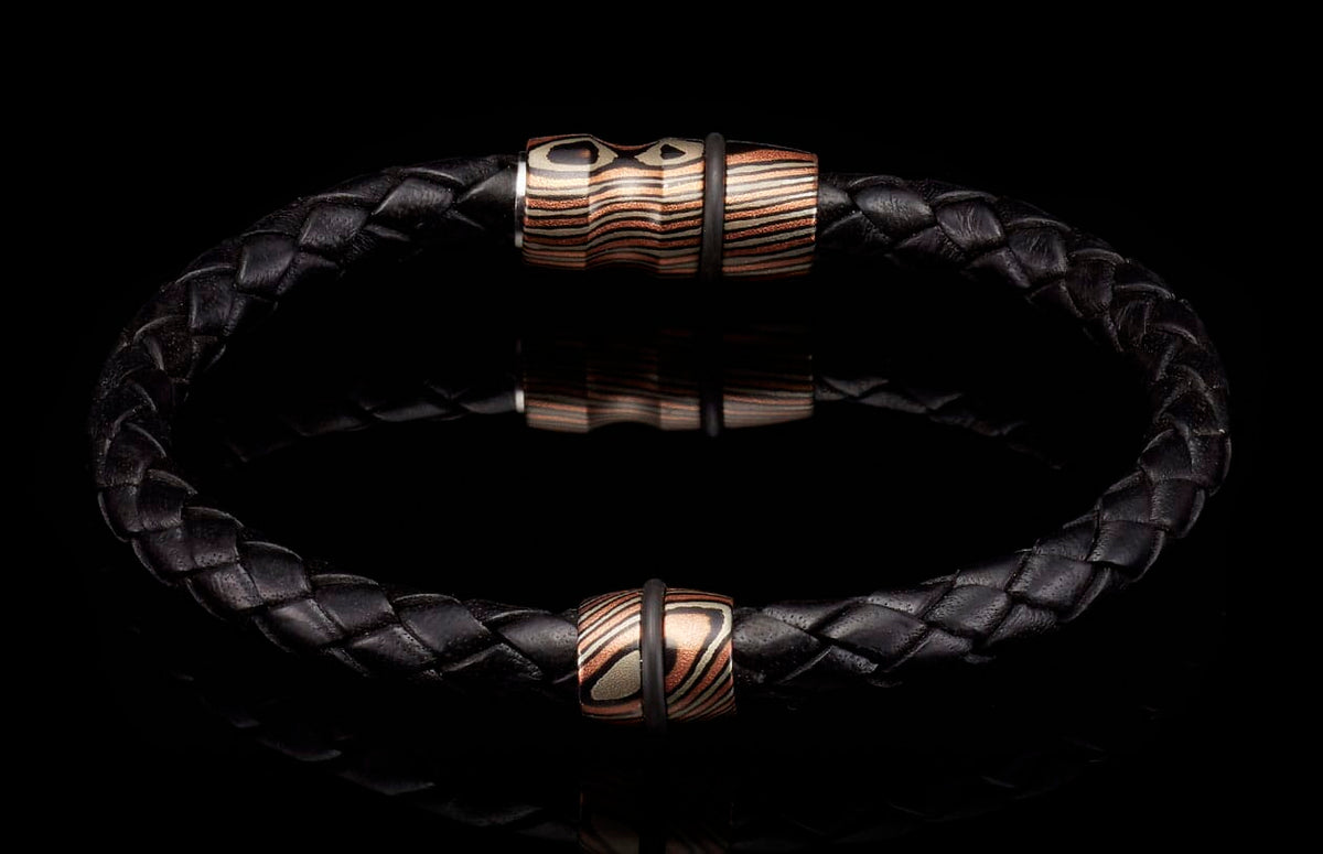 Black braided leather bracelet with two striped copper, silver, and black beads, each with a black rubber ring, on a reflective surface.