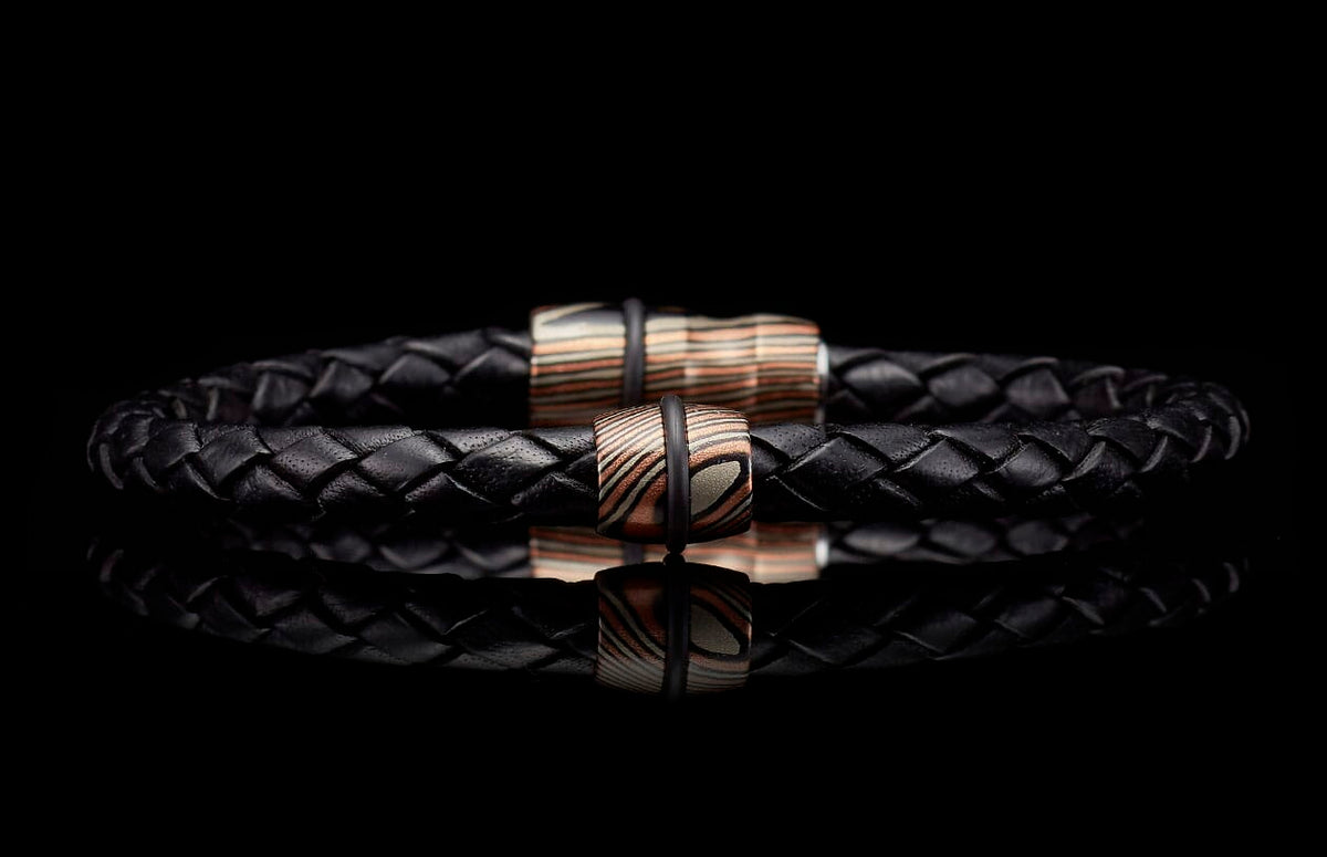 Black braided leather bracelet with two cylindrical copper and silver patterned beads, reflected on a dark surface.