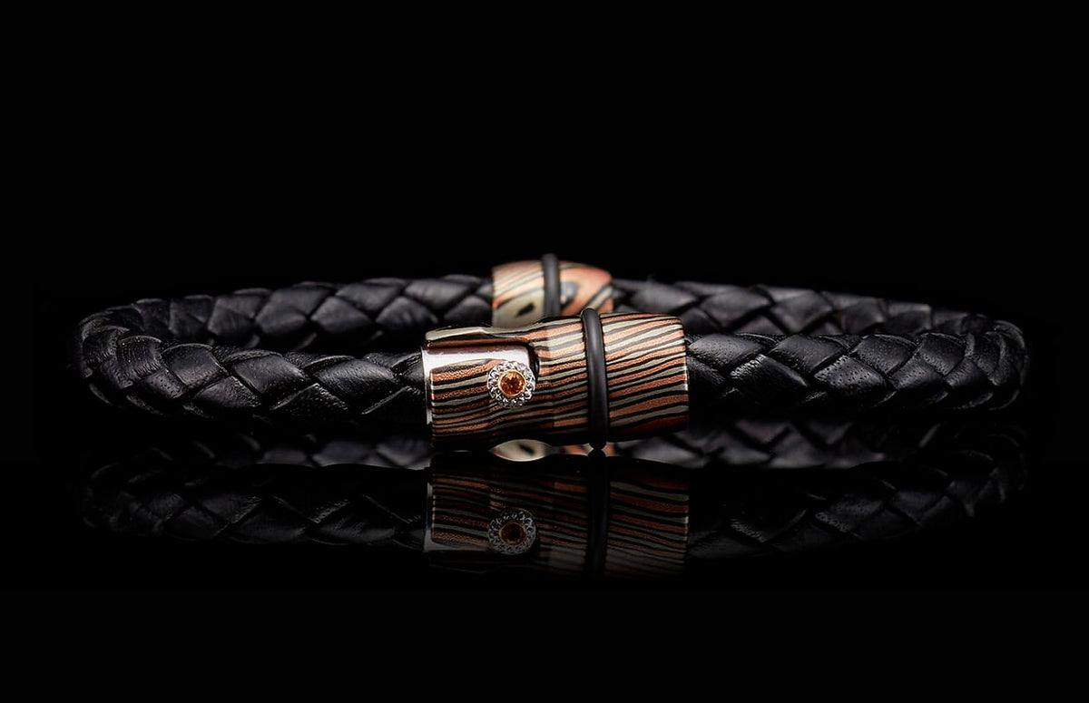 Black braided leather bracelet with a striped metallic clasp in bronze and silver tones, featuring an orange gemstone.