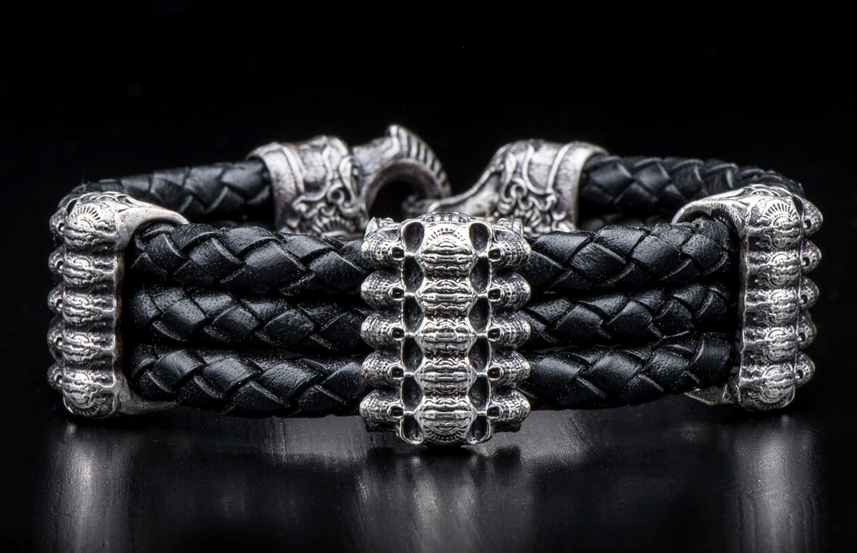 Black braided leather bracelet with silver skull-like center bead and ornate silver end caps, featuring a hook clasp.
