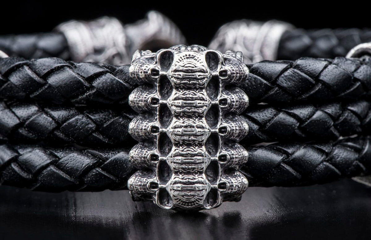 Black braided leather bracelet with an intricate silver-toned metal centerpiece featuring a stacked skull motif, resting on a dark surface.