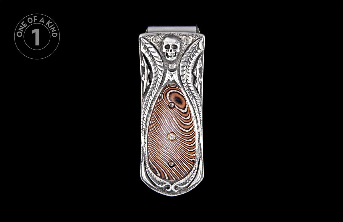 Silver money clip with an embossed skull, ornate carvings, and a brown Damascus patterned inlay with two gems.