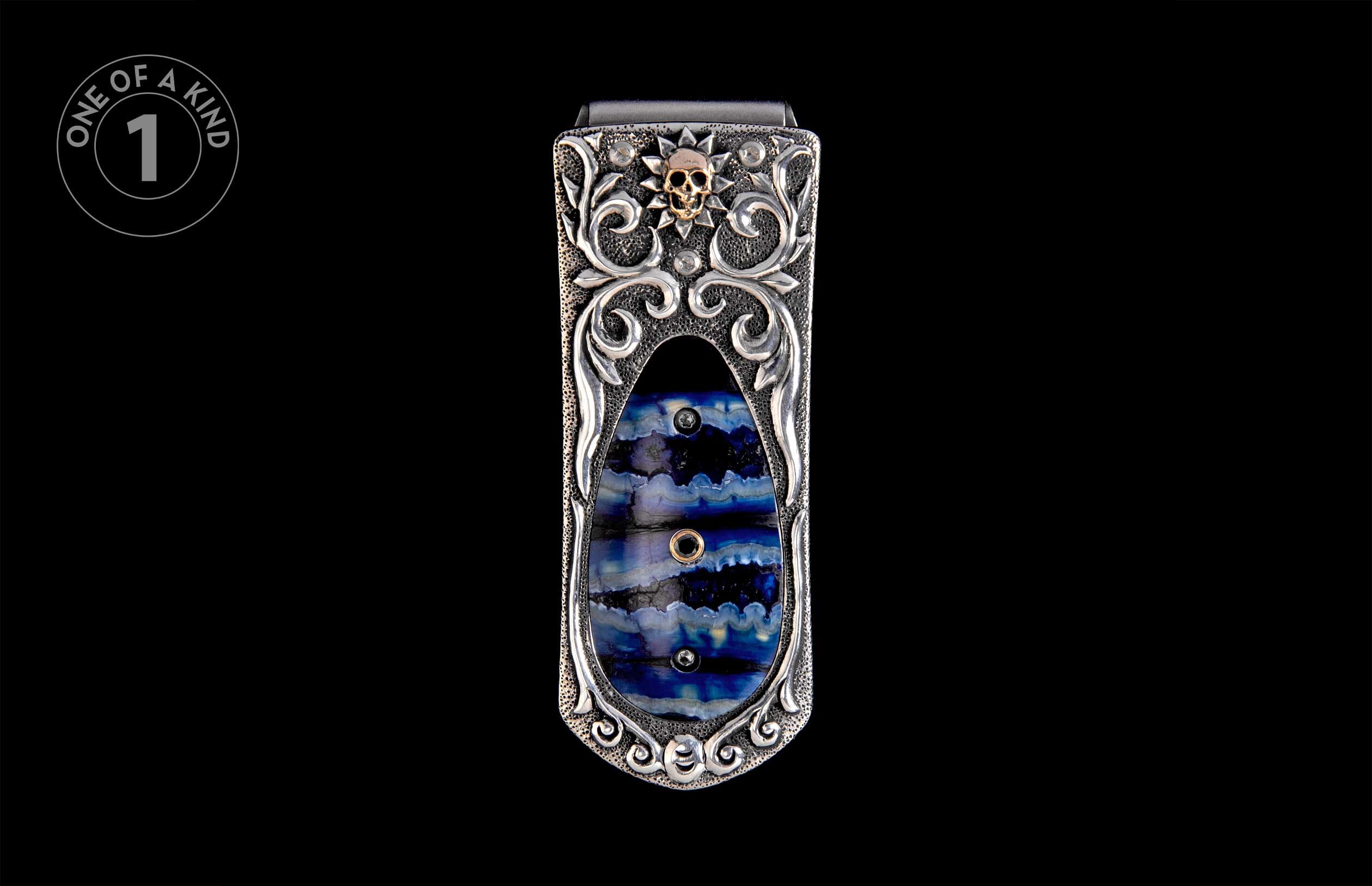 Ornate silver money clip with a gold skull, sunburst, scrollwork, and a layered blue stone inlay.