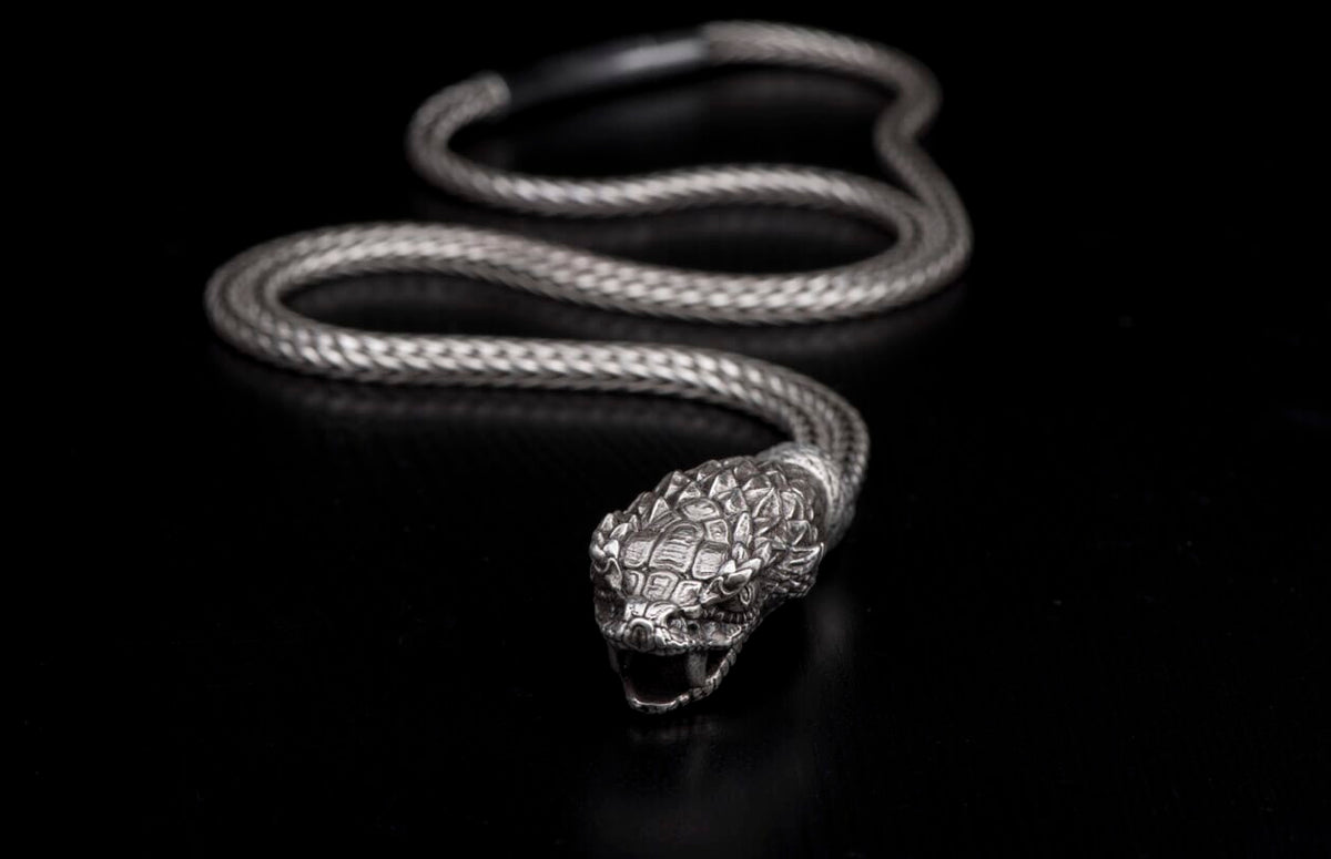 Silver snake-shaped jewelry with a braided body and detailed head with open jaws, on a black background.