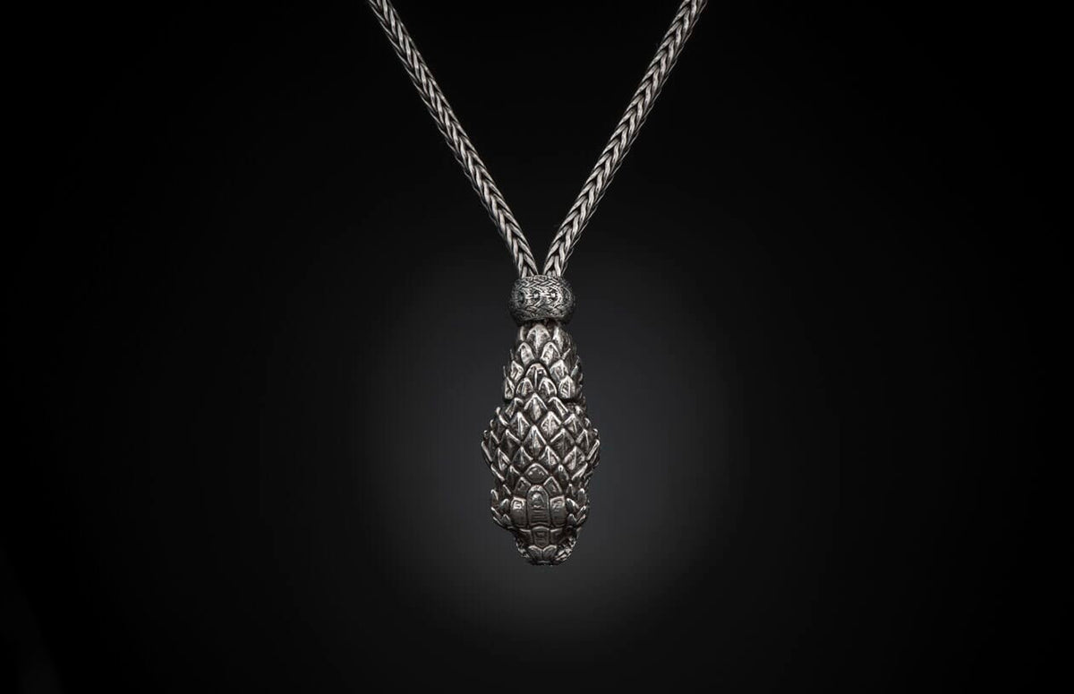 Silver braided necklace with a textured, tapered pendant featuring overlapping scale-like elements, against a black background.