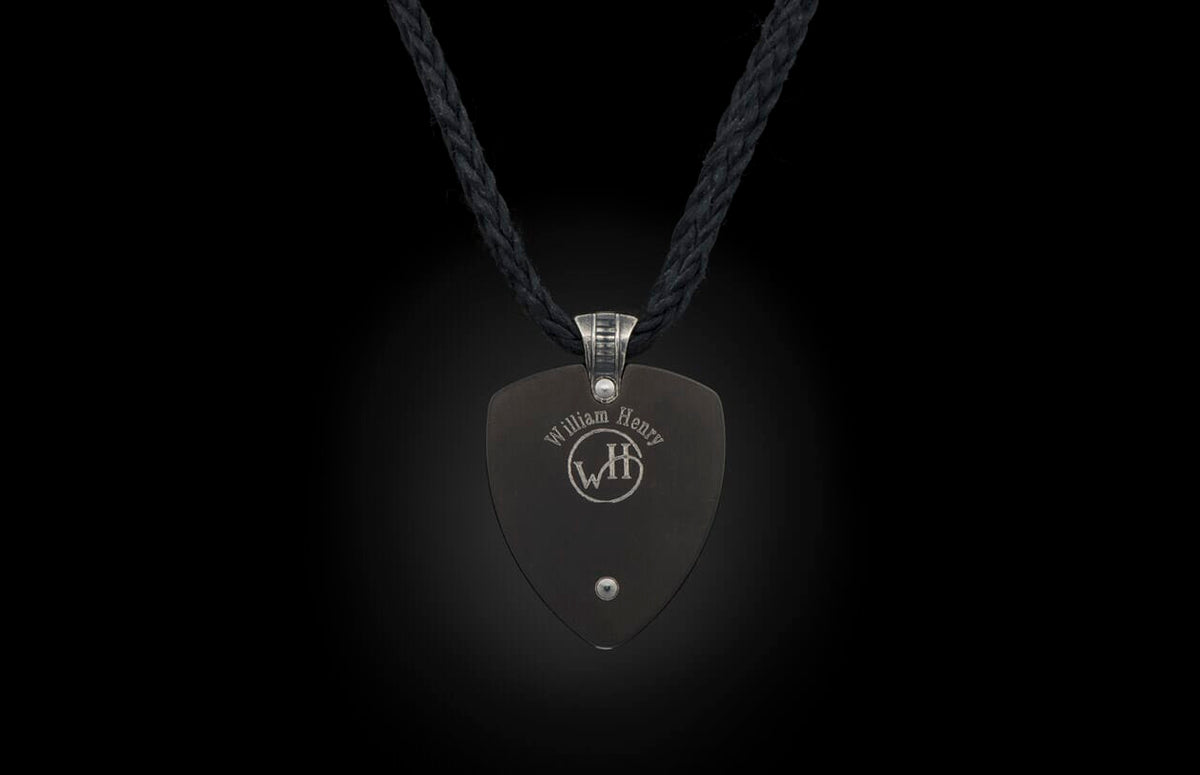 A dark grey pendant by William Henry, shield-shaped, with a silver rivet and etched logo, on a black braided cord.