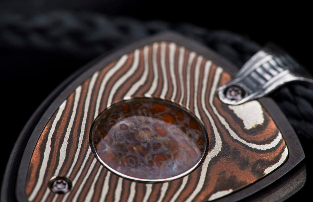 Decorative jewelry pendant featuring patterned copper and silver metal with a central mottled reddish-brown stone and ribbed silver bail on a black cord.