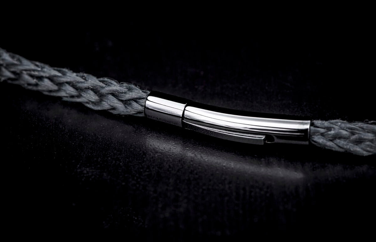 A dark grey braided bracelet with a sleek, polished silver-tone clasp on a dark textured surface.