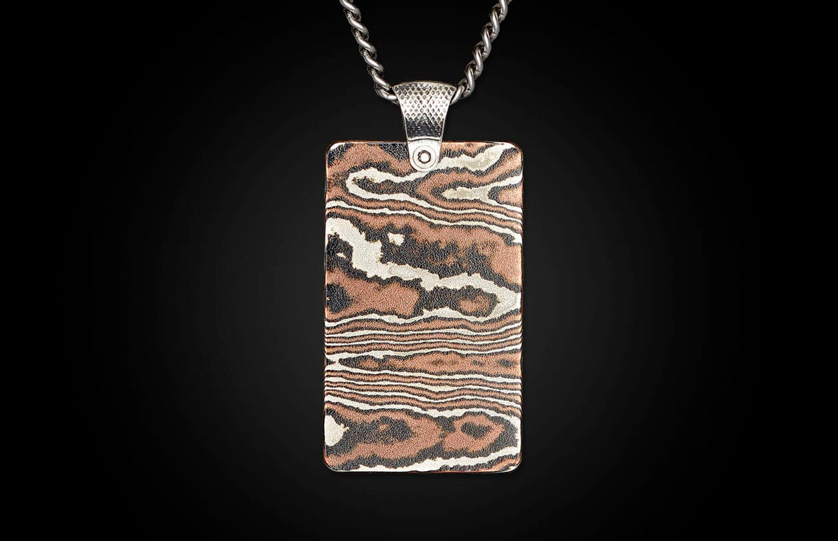 Rectangular metal pendant with a silver chain and bail, featuring a swirling woodgrain pattern in copper, silver, and dark tones.