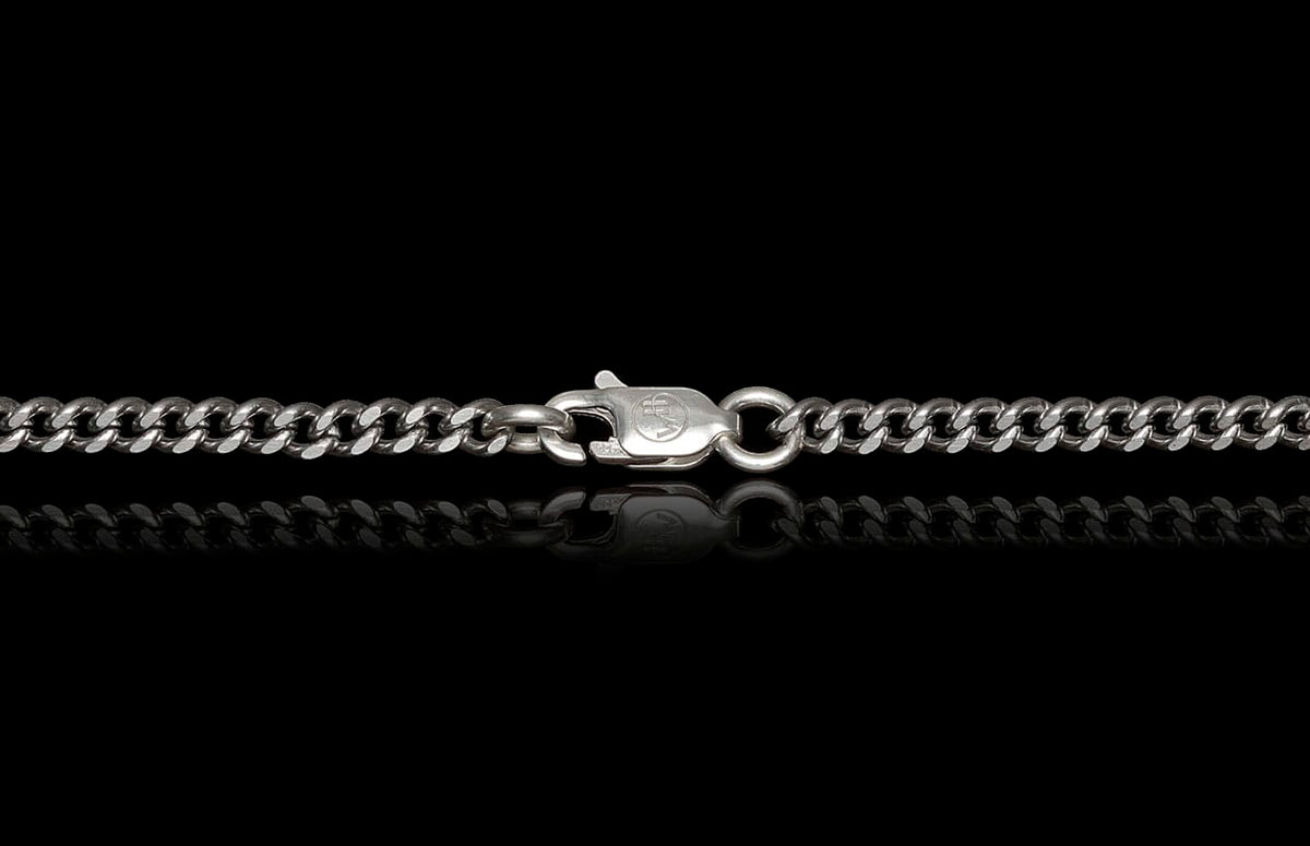Silver curb chain by Mokume Forge with a lobster clasp featuring a circular emblem, reflected on black.