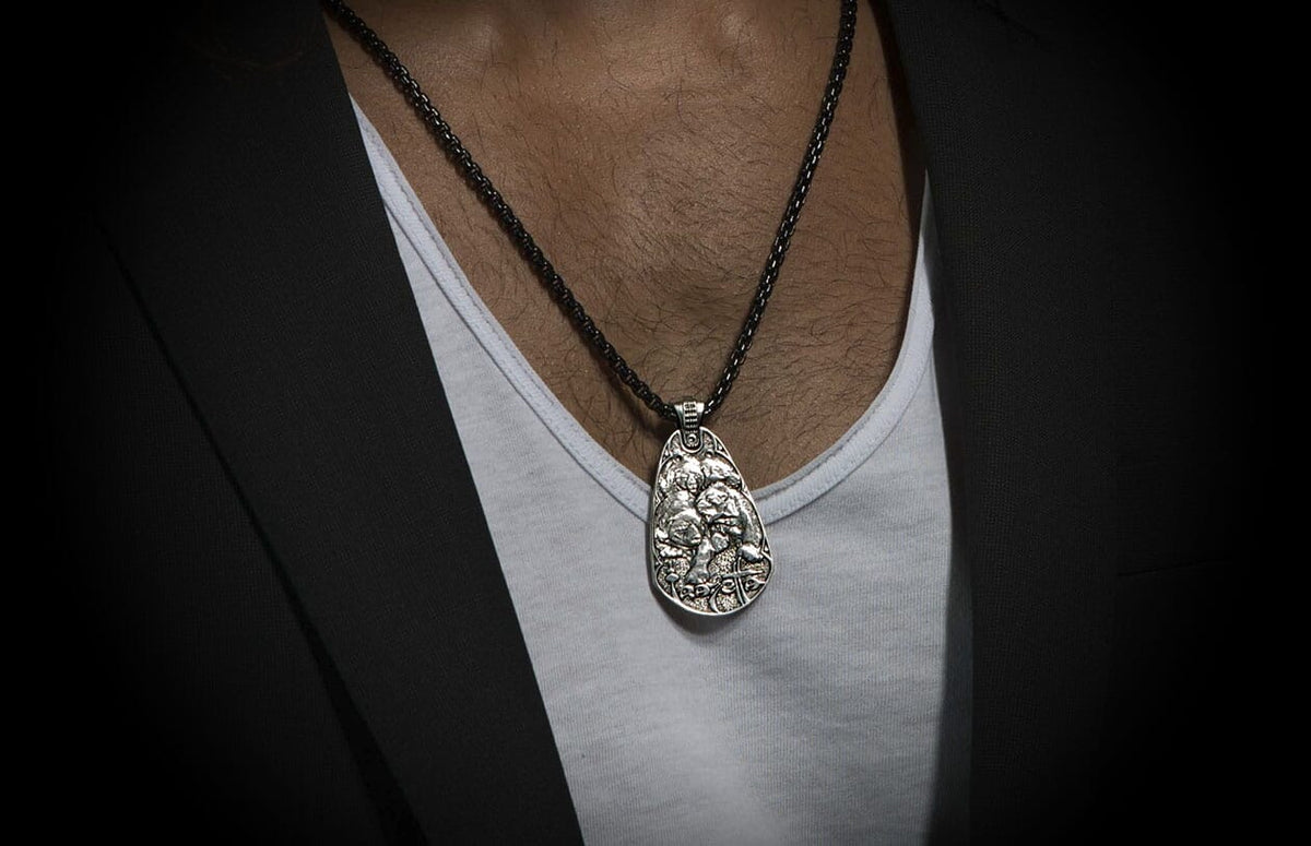 Silver Guardians pendant necklace with detailed relief carving, on a dark textured chain, worn over a white undershirt and black blazer.